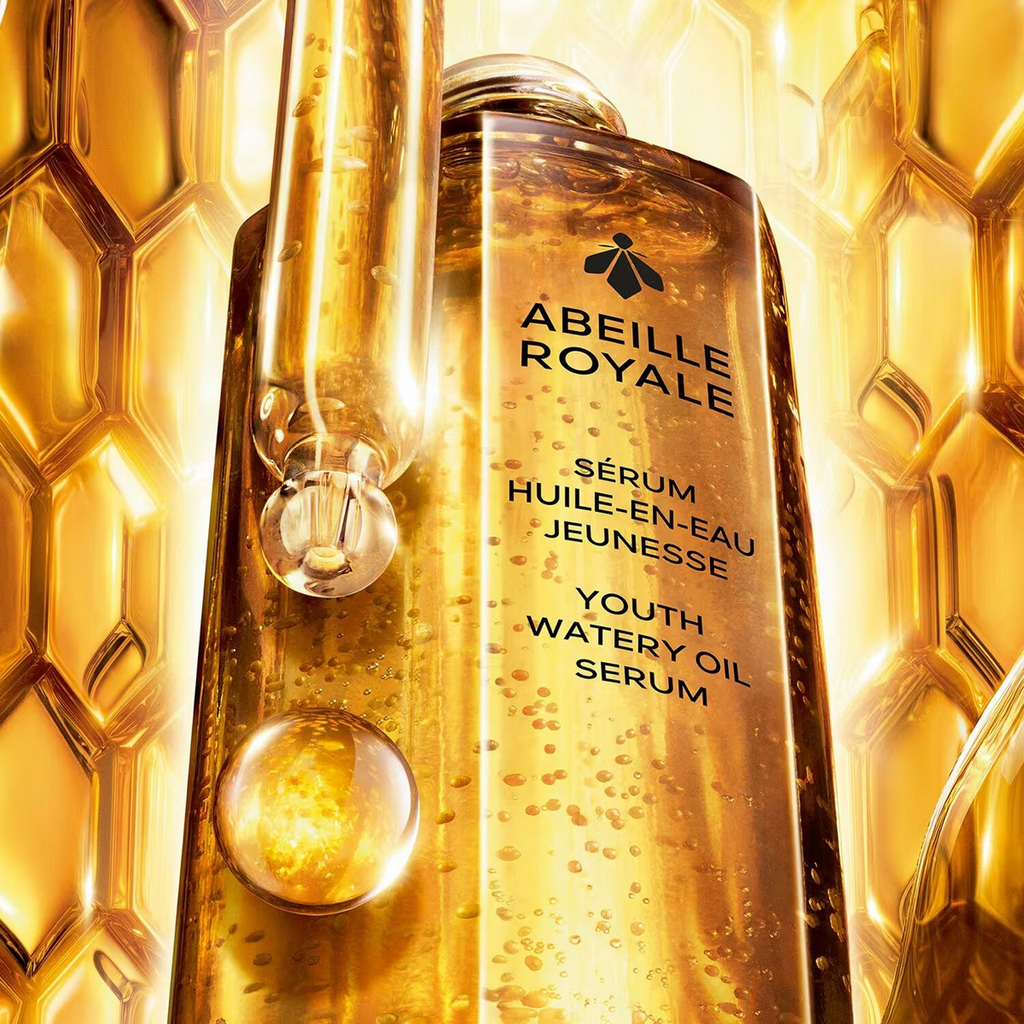 Close-up image of the Guerlain Abeille Royale Youth Watery Oil Serum bottle with golden microbeads and a glass dropper, set against a glowing honeycomb background